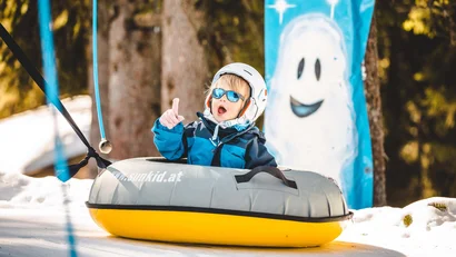 Kind in Helm und Sonnenbrille auf Schneereifenrutsche im Winterwald