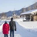 Zwei Personen gehen im Schneeweg an einer verschneiten Holzhütte vorbei