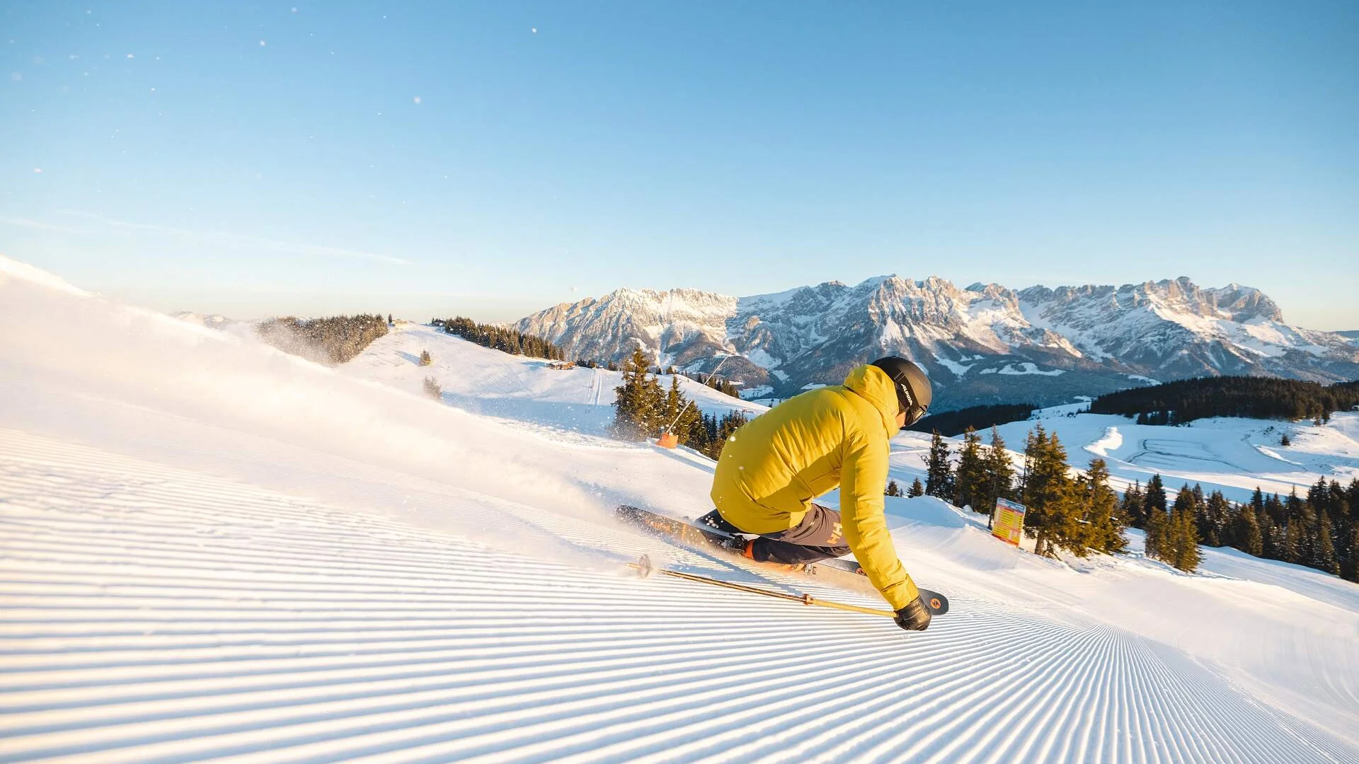 Skifahrer in gelber Jacke fährt auf präparierter Piste mit Bergkulisse