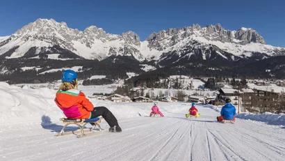 Menschen rodeln auf einem Schneehang vor verschneiten Bergen bei klarem Himmel